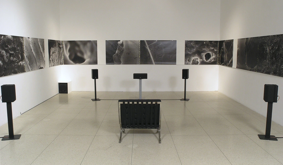 Image de l'œuvre Sweeping Spirals, par AE. Installation composée de haut-parleurs, de photographies accrochées au mur et d'une chaise. Réalisée de 2003 à 2006.