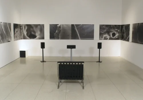 Image de l'œuvre Sweeping Spirals, par AE. Installation composée de haut-parleurs, de photographies accrochées au mur et d'une chaise. Réalisée entre 2003-2006.
