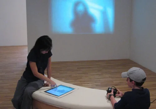 Deux spectateurs interagissent avec une tablette et une caméra, disposées dans l'œuvre Digitale, par Alexandre Castonguay. Installation et vidéo projection. Réalisée entre 2003-2004. Dans le cadre de l'exposition À l’intérieur/Inside, durant la 3rd Beijing International New Media Arts Exhibition and Symposium.